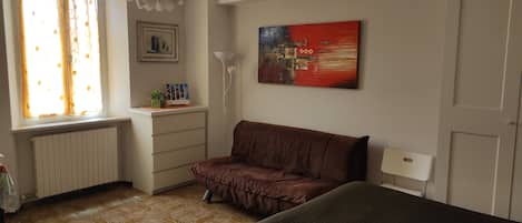 A bedroom with a bed, a brown sofa, a window with curtains, a radiator, a desk with a plant, a framed picture, and a painting on the wall.