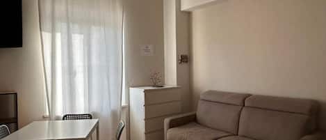 A living room with a beige sofa, a dining table with chairs, a TV mounted on the wall, and a window with curtains.