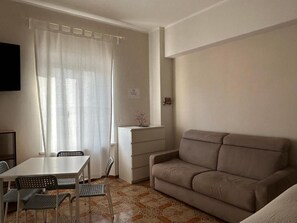 Una sala de estar con un sofá color crema, una mesa de comedor con sillas, un televisor montado en la pared y un ventanal con cortinas.