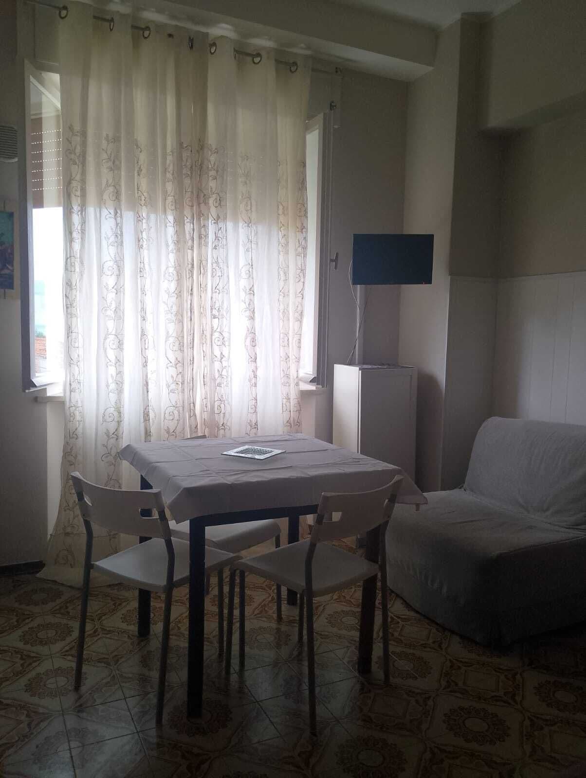Una habitación con una mesa de comedor, dos sillas, un sofá, un televisor de pantalla plana y un ventanal con cortinas.