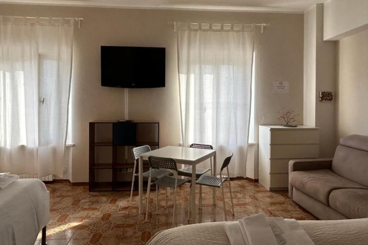 A hotel room with a bed, a sofa, a dining table with chairs, a TV mounted on the wall, and a small cabinet.
