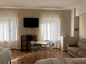 A hotel room with a bed, a sofa, a dining table with chairs, a TV mounted on the wall, and a small cabinet.