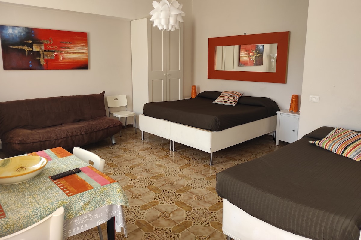 A bedroom with a bed, a sofa, a small table with a bowl and remote, and a mirror on the wall.