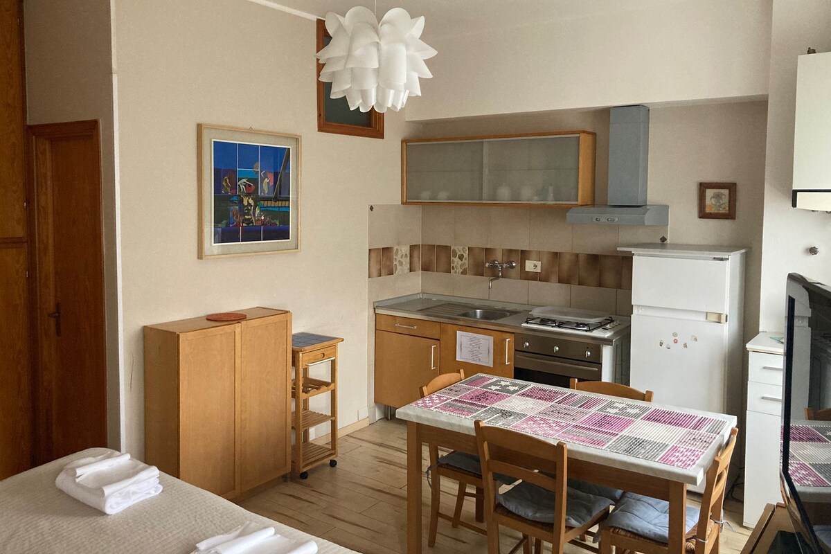 A compact kitchen with a stove, sink, and refrigerator. There is a dining table with chairs and a small wooden cabinet. A framed picture hangs on the wall above the sink.
