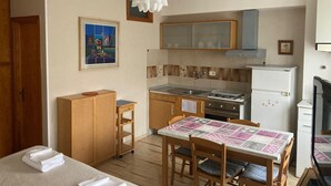 A compact kitchen with a stove, sink, and refrigerator. There is a dining table with chairs and a small wooden cabinet. A framed picture hangs on the wall above the sink.