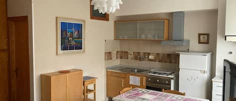 Cocina compacta con estufa, lavaplatos y refrigerador. Cuenta con una mesa de comedor con sillas y un pequeño armario de madera. Sobre el lavaplatos, hay un cuadro enmarcado.