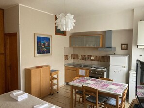 A compact kitchen with a stove, sink, and refrigerator. There is a dining table with chairs and a small wooden cabinet. A framed picture hangs on the wall above the sink.