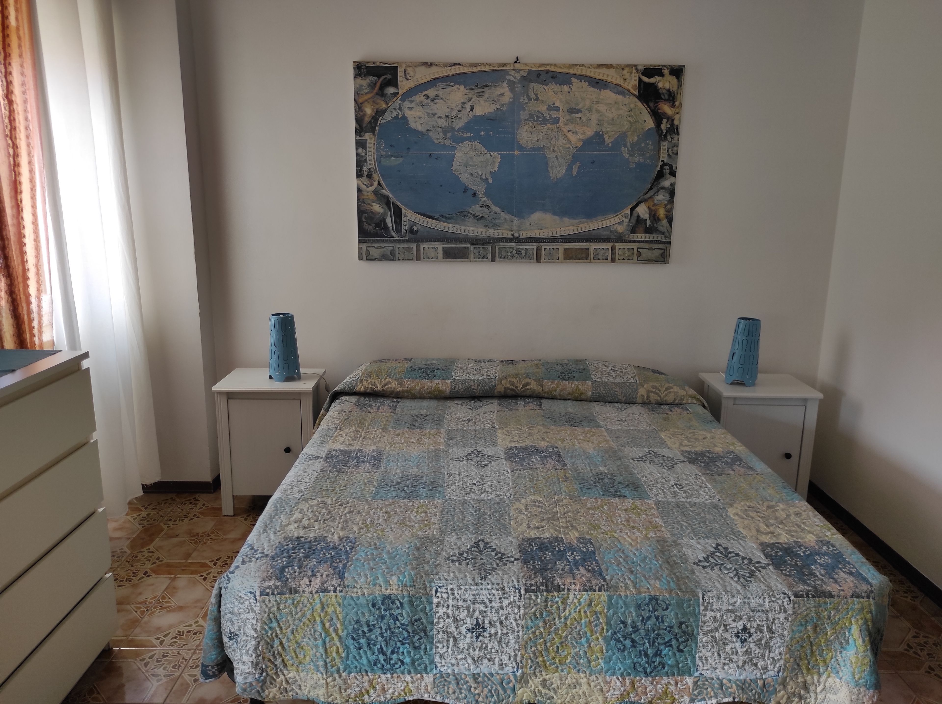 A bedroom with a large world map on the wall, two bedside tables with vases, a bed with a patterned quilt, and a dresser with drawers.