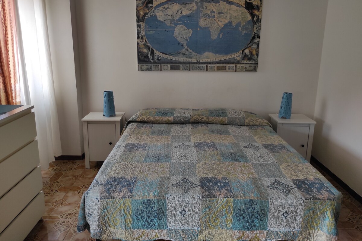 A bedroom with a large world map on the wall, two bedside tables with vases, a bed with a patterned quilt, and a dresser with drawers.