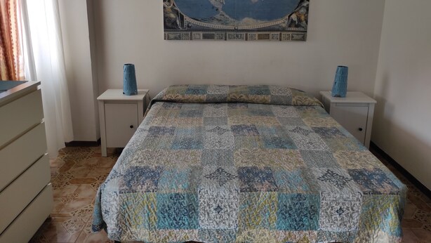 A bedroom with a large world map on the wall, two bedside tables with vases, a bed with a patterned quilt, and a dresser with drawers.