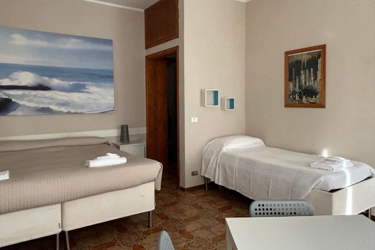 A hotel room with two beds, a painting of a beach scene, a small table, and a chair.