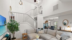 A spacious living area with a large sectional sofa, a small round coffee table, and a potted plant. A staircase with white railings leads to an upper level.