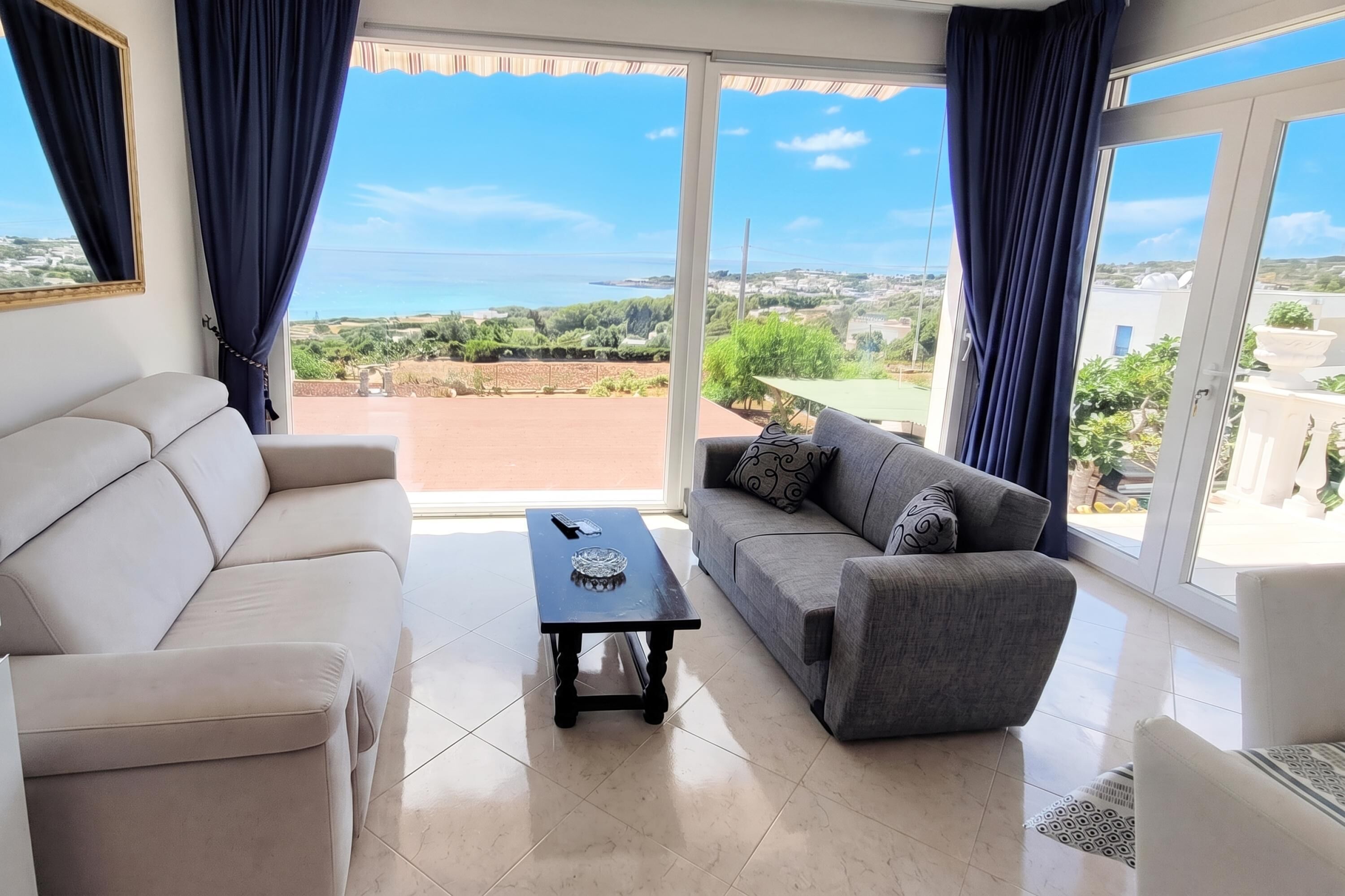 A living room with a beige sofa, a grey sofa with patterned cushions, and a small black table with a glass bowl. Large windows offer a view of a green landscape and buildings.