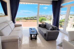 Sala de estar con un sofá beige, un sofá gris con cojines estampados y una pequeña mesa negra con un cuenco de vidrio. Amplias ventanas ofrecen vistas a un paisaje verde y edificios.
