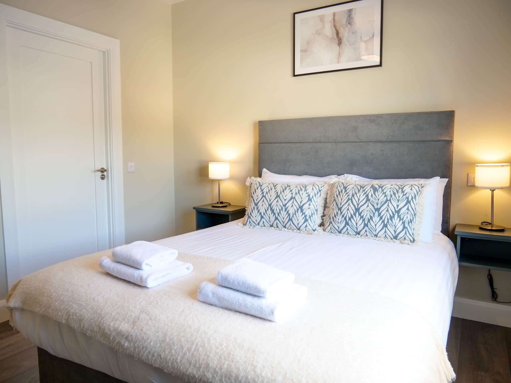 A neatly made bed with white linens and folded towels, a grey upholstered headboard, and two bedside lamps on matching nightstands.