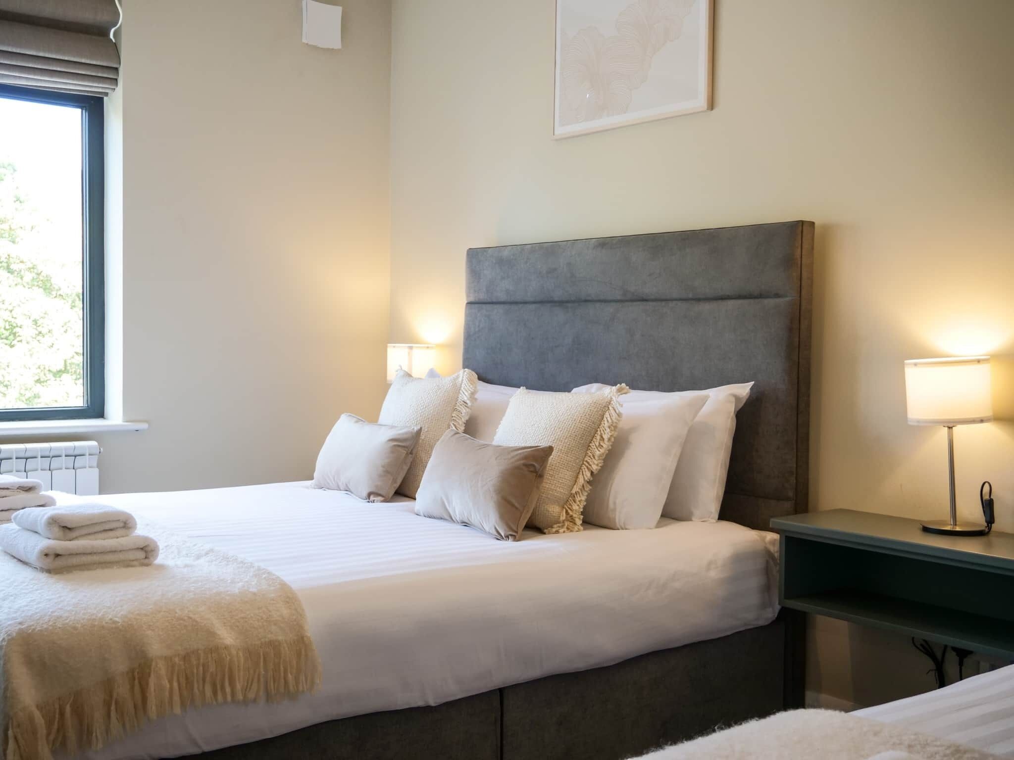 Ein ordentlich bezogenes Bett mit weißen Leinentüchern und einer beigefarbenen Tagesdecke, ein Nachttisch mit einer Lampe und ein Fenster mit Blick auf Grünflächen.