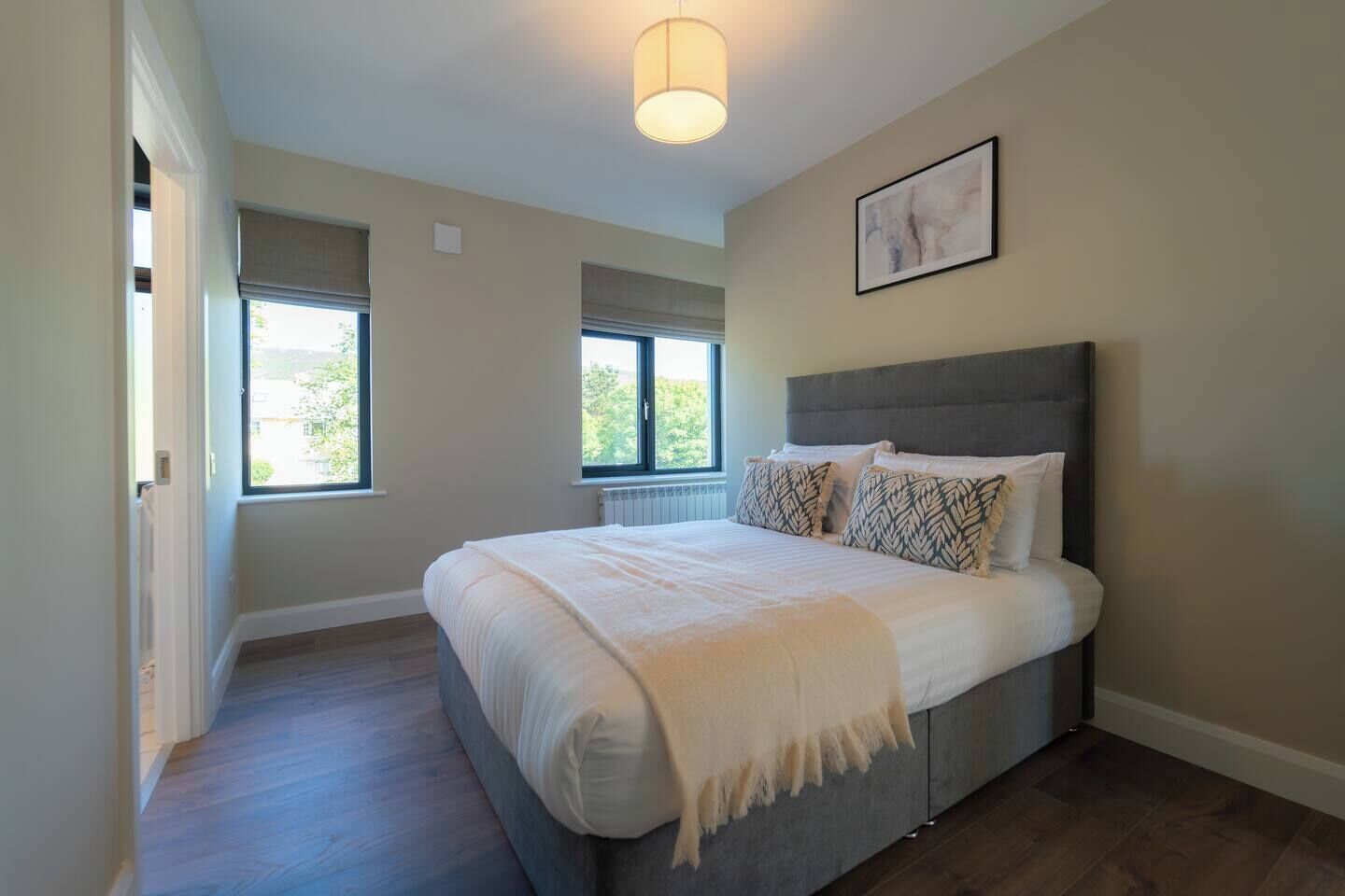 Une chambre à coucher avec un grand lit, deux fenêtres, un luminaire au plafond et un tableau encadré au mur.
