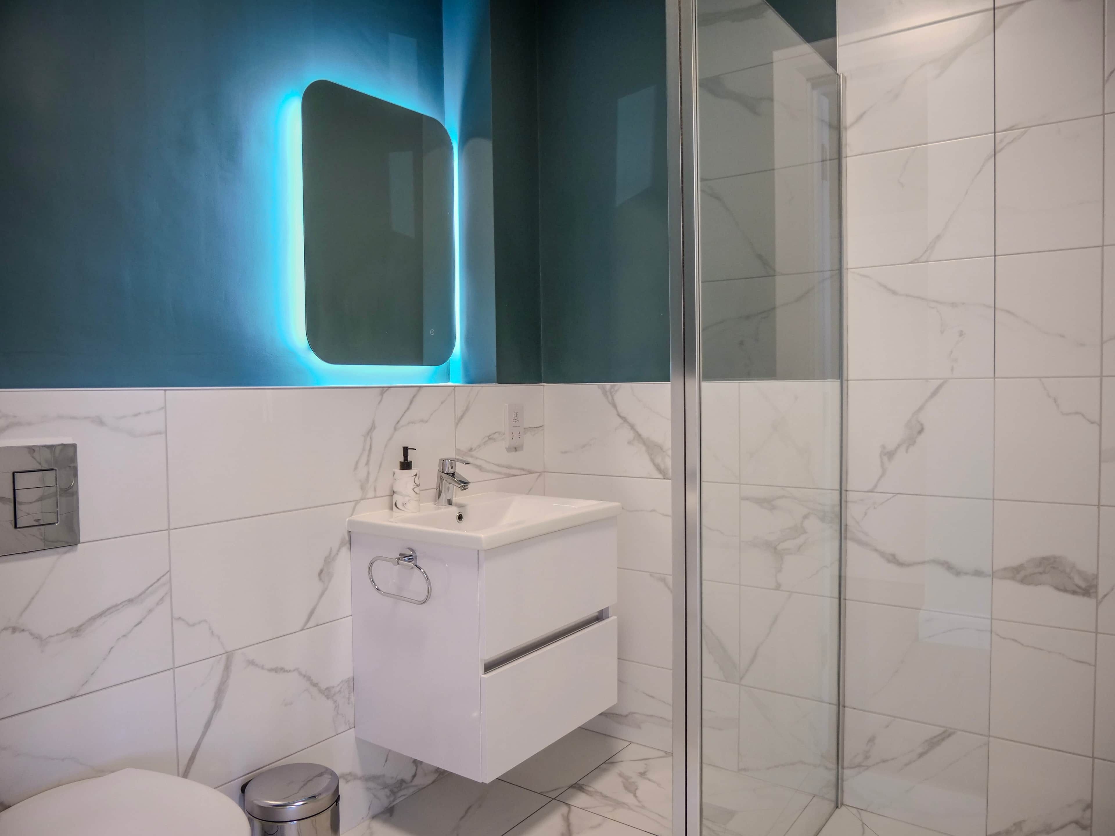 A modern bathroom with a glass shower enclosure, white marble tiles, and a built-in sink with a soap dispenser.