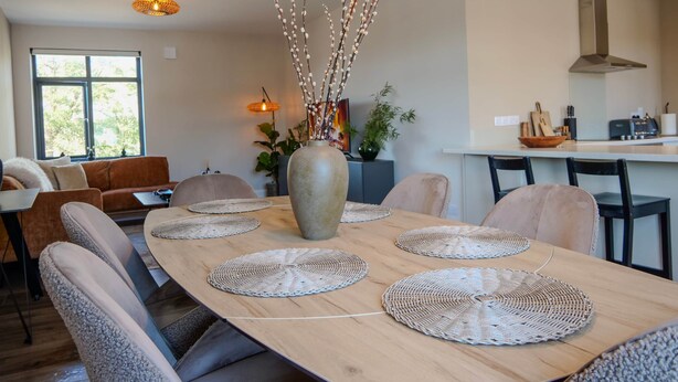 Un espace repas moderne avec une table en bois, des sets de table en fibres tressées et un vase décoré de branches.