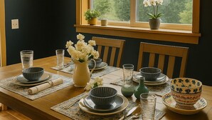 Een eethoek met een houten tafel voor vier personen, omringd door stoelen. Boven de tafel hangen twee pendelverlichting, en een raam met uitzicht op bomen.