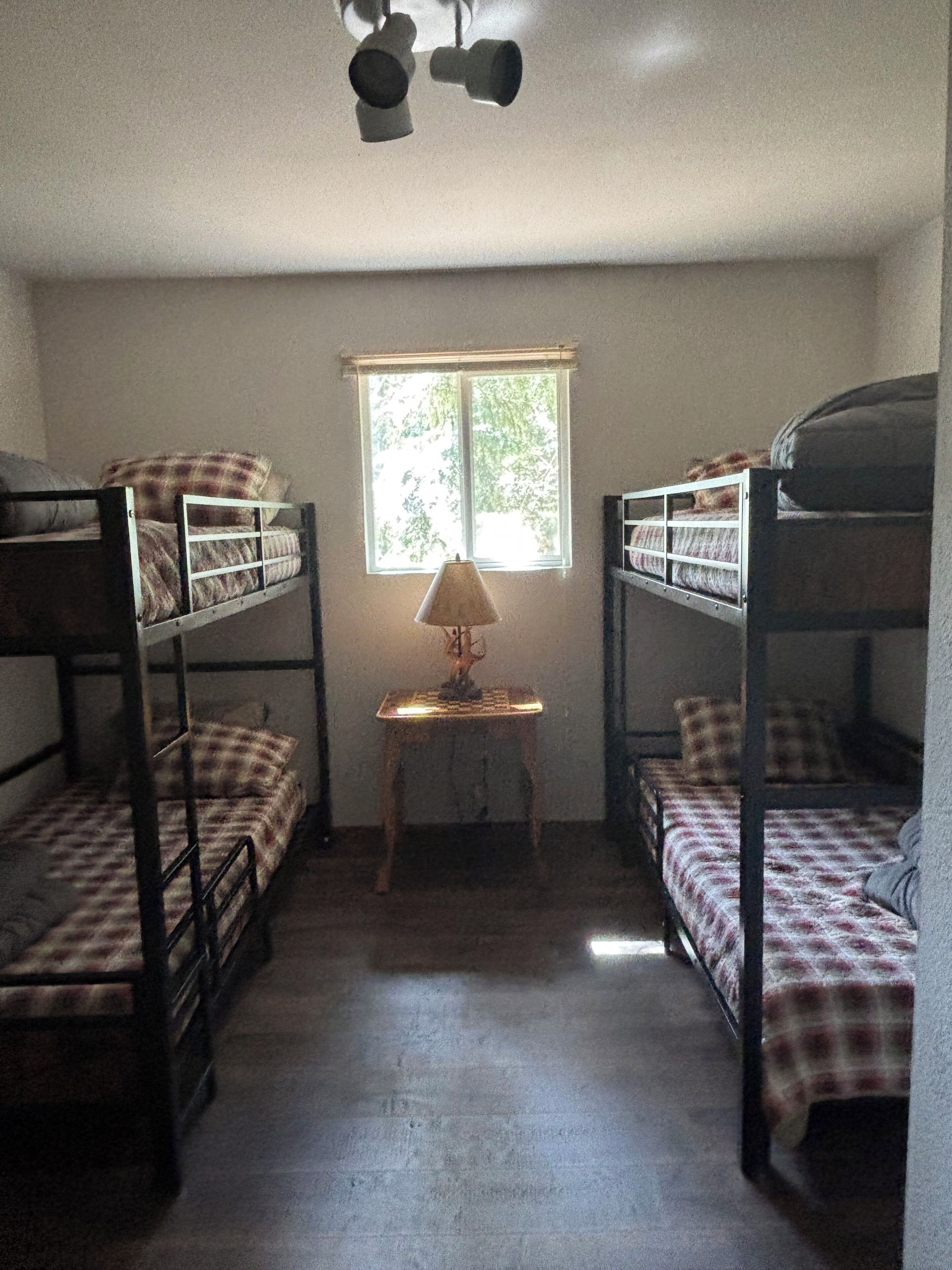A dormitory room with bunk beds, a small table with a lamp, and a window.
