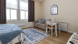 A cozy room with a bed, a desk, and a chair. There is a window with curtains, a radiator, and a small table with a lamp.