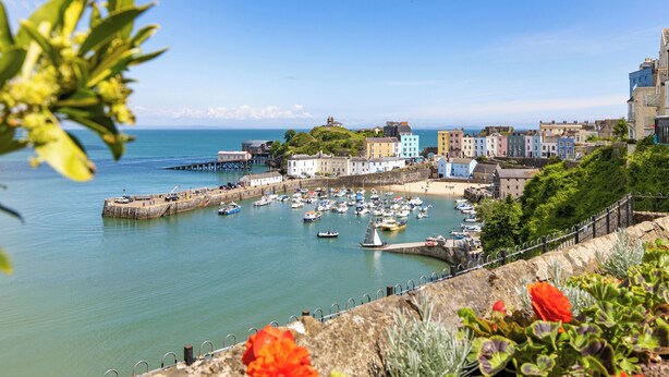 A coastal town with a harbor filled with boats, colorful buildings, and a prominent hill with a structure on top.