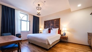 A hotel room with a bed, a desk, a chair, a nightstand with lamps, and a window with curtains. - Agarani Estate (Telavi)