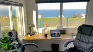 A room with a desk, two chairs, a laptop, a lamp, and a view of the sea.