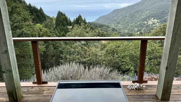 A wooden deck with a metal table, overlooking a forested hillside and a body of water. - Stunning Ocean View Getaway- Private and Serene (Whitethorn)