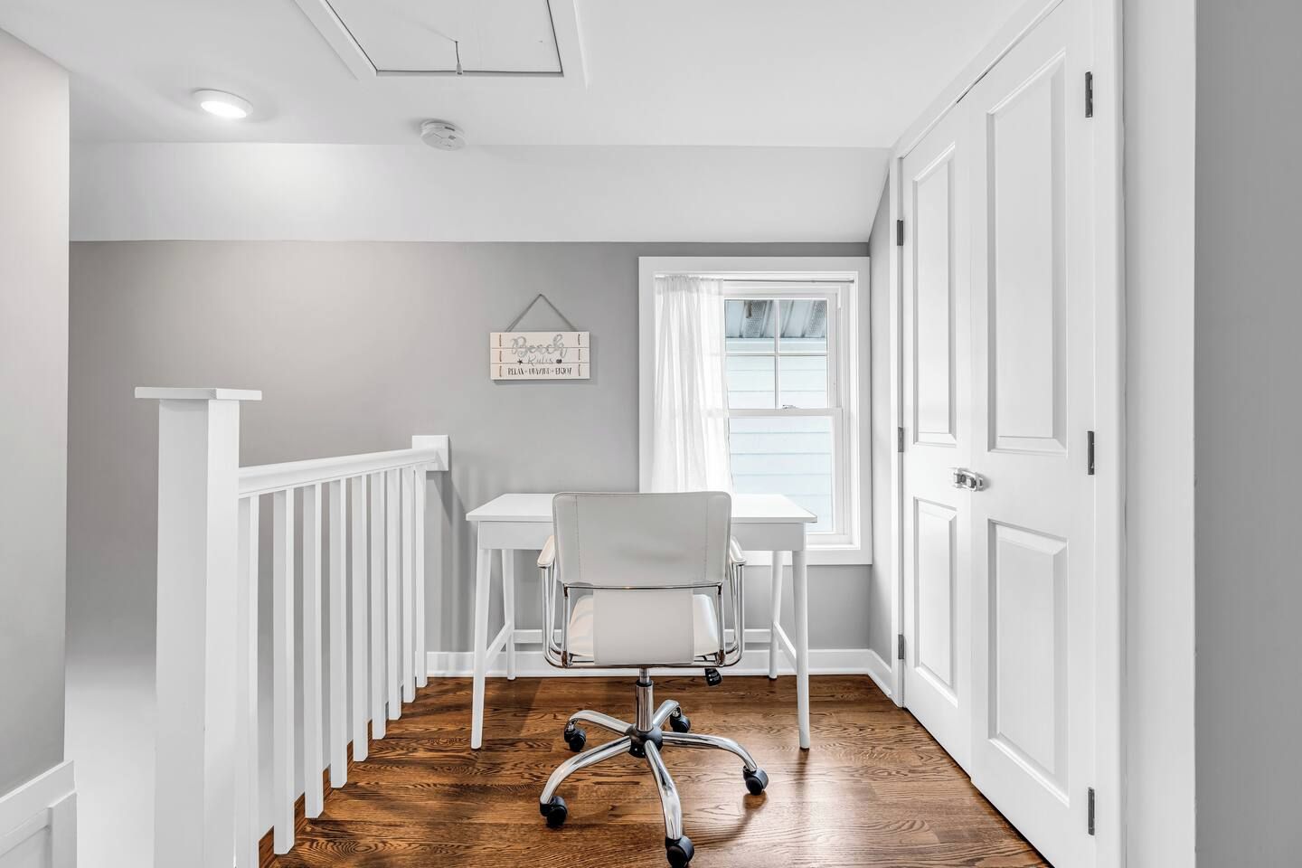 A narrow hallway with a white railing, a desk with a chair, a window with curtains, and a door.