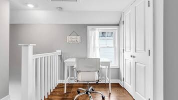 A narrow hallway with a white railing, a desk with a chair, a window with curtains, and a door.
