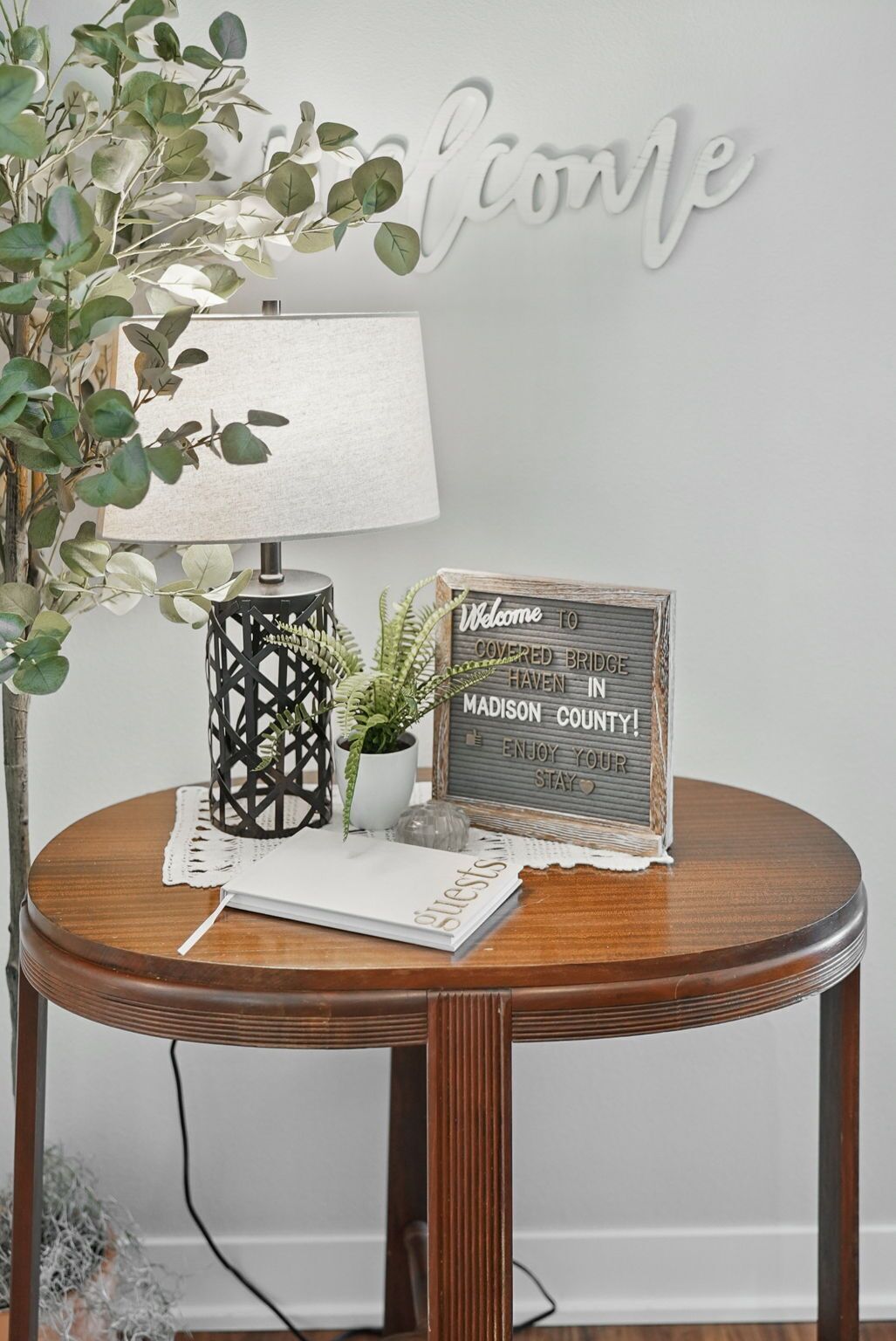 A round wooden table with a lamp, a welcome sign, and a small potted plant.