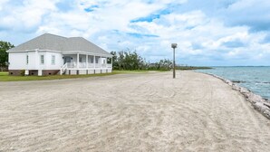 A white building with a porch, a sandy beach, and a body of water in the background. - Welcome to Core Sound Retreat – Your Peaceful Down East Getaway (Davis)