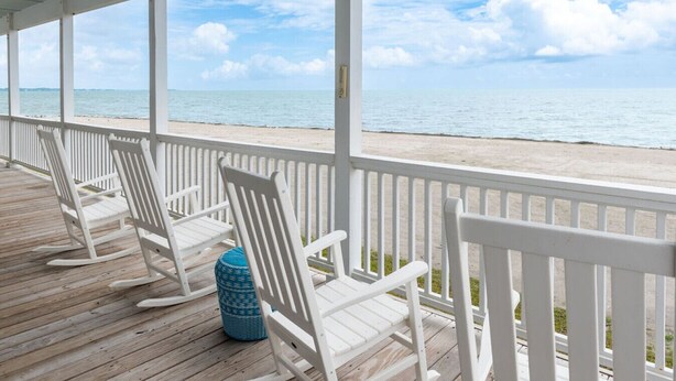 A covered wooden deck with white rocking chairs and a blue patterned ottoman. - Welcome to Core Sound Retreat – Your Peaceful Down East Getaway (Davis)