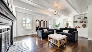 A spacious living room with dark wood flooring, white walls, and black leather sofas. There is a wooden bench and a ceiling light fixture.