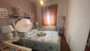Ein Schlafzimmer mit Bett, Nachttischen, einer Lampe, einer Wanduhr, einem Fenster mit Vorhängen und einer Heizung.