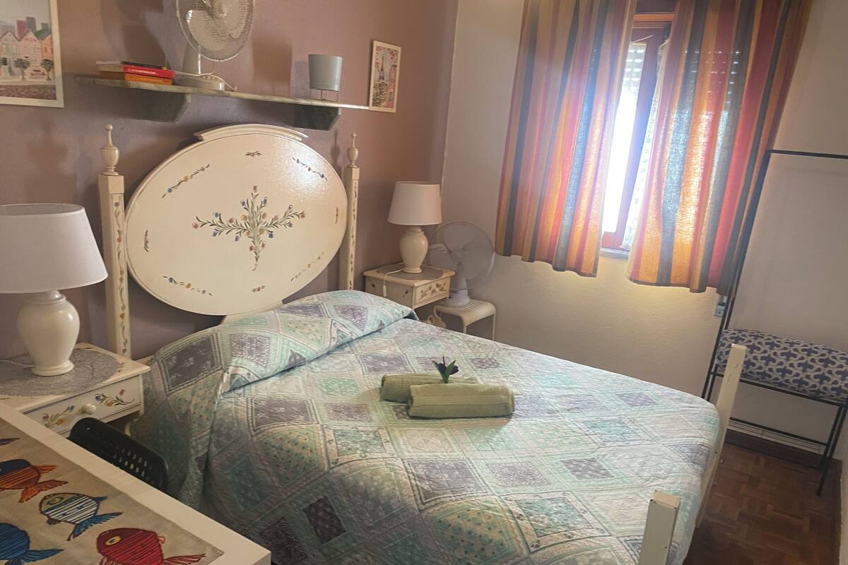 A bedroom with a bed, bedside tables, a lamp, a fan, and a window with curtains.