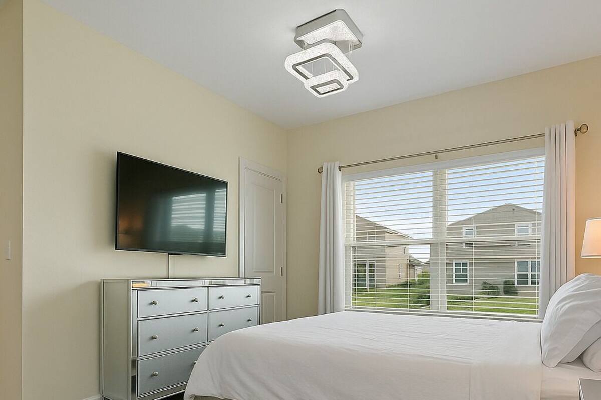 A bedroom with a bed, a dresser, a television, and a window with blinds.