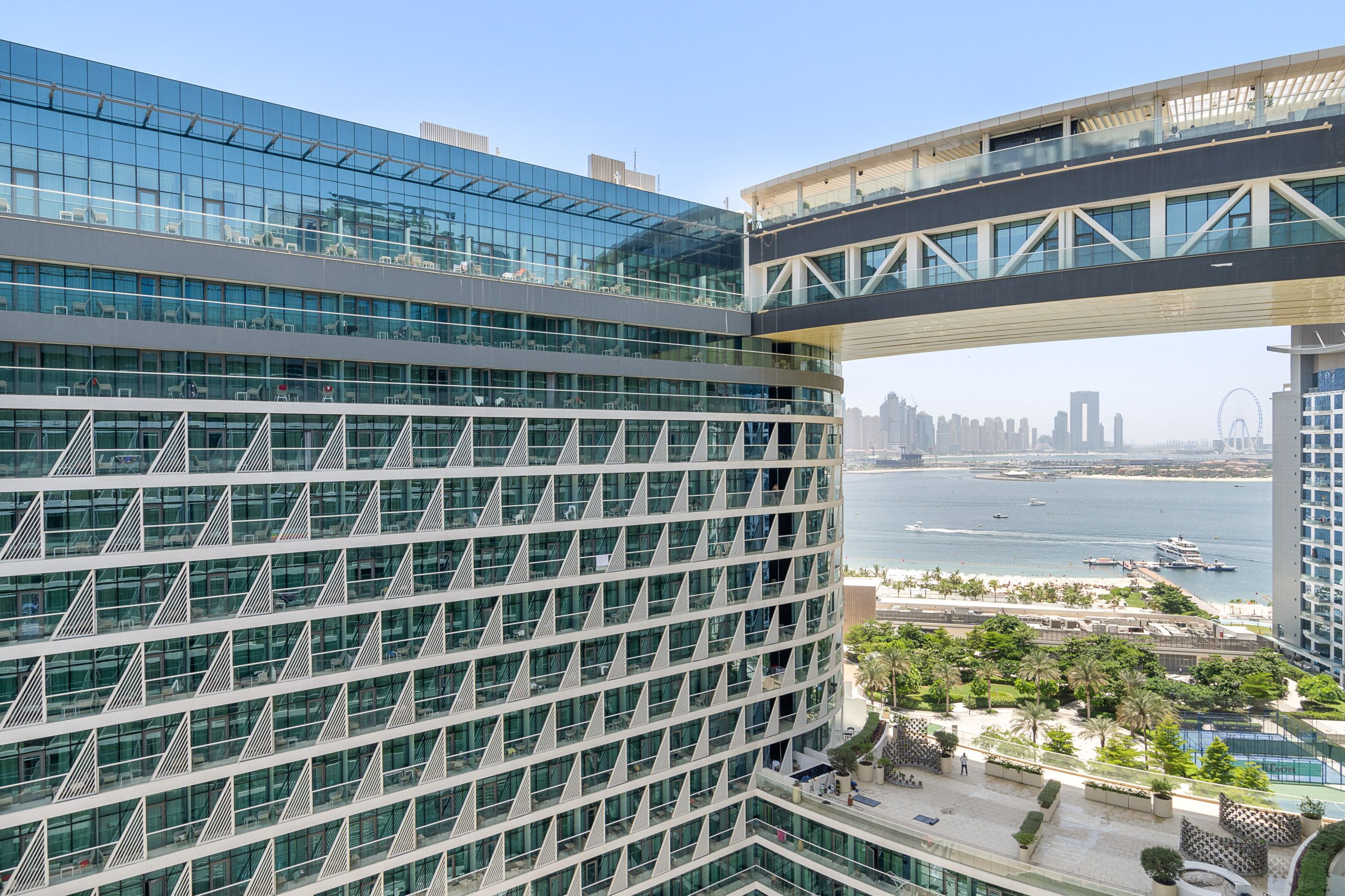 Un edificio de gran altura con fachada de vidrio, vistas al agua y al horizonte de la ciudad, y un puente peatonal.