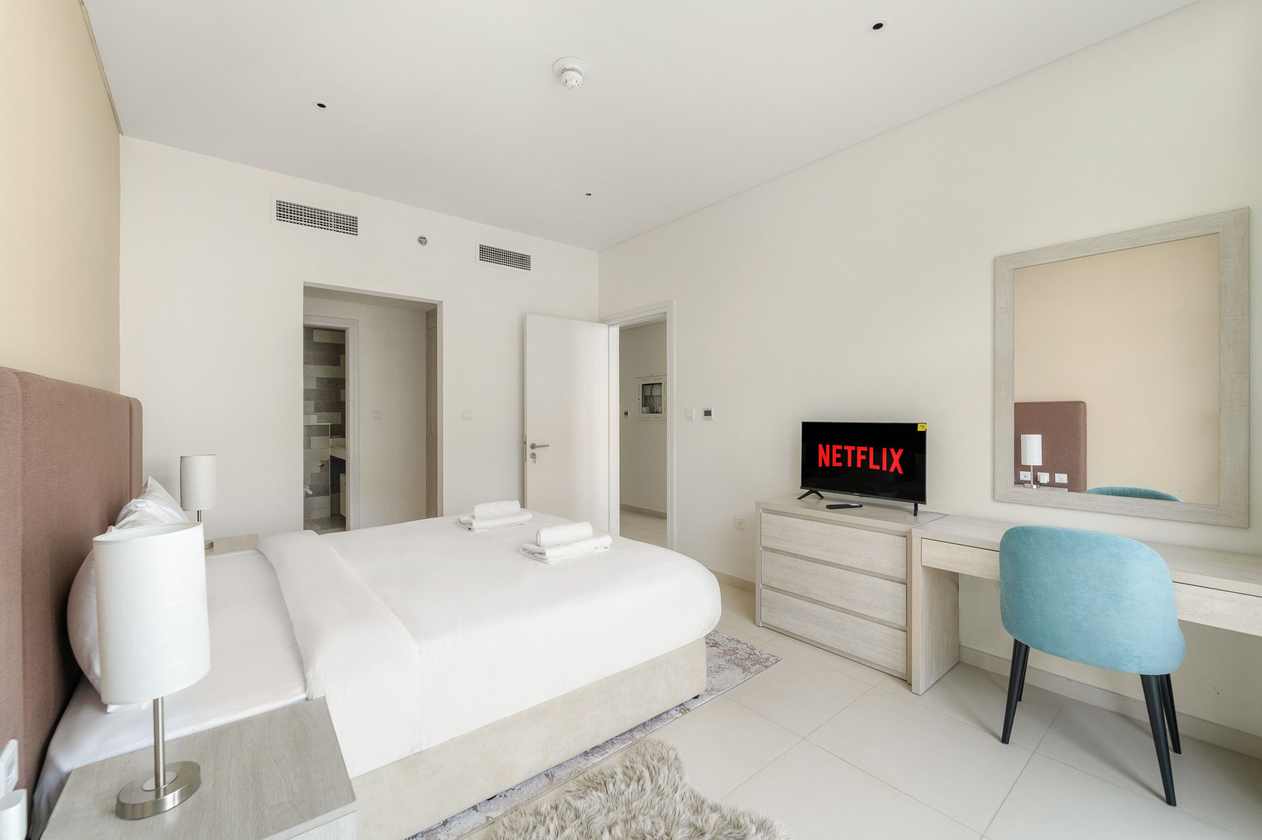 A modern hotel room with a large bed, a desk, a chair, a TV displaying Netflix, and a mirror.