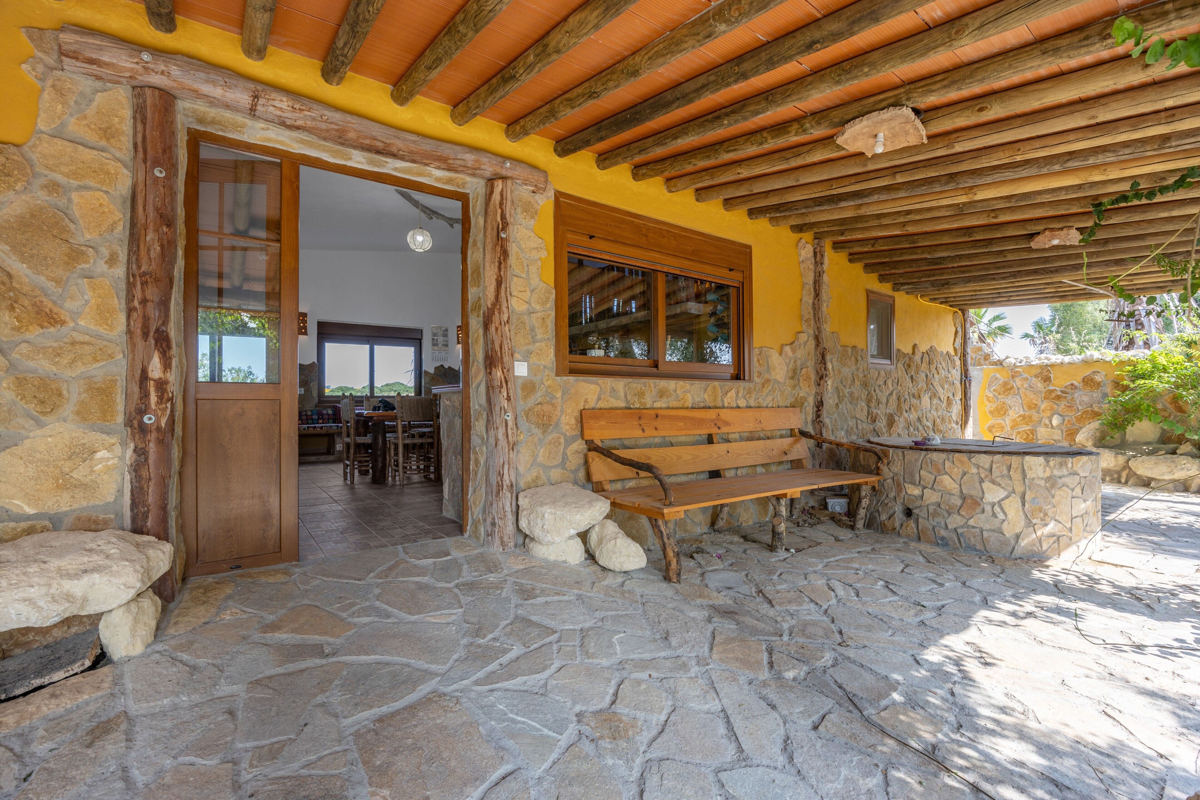 A stone-floored patio with a wooden bench and a stone table, a wooden door, and a yellow wall with a window.