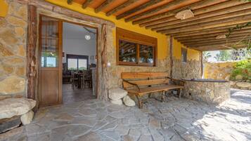 A stone-floored patio with a wooden bench and a stone table, a wooden door, and a yellow wall with a window.