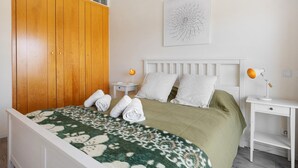 Un dormitorio con cama, un armario de madera, una mesita de noche blanca con lámpara y una obra de arte en la pared.