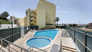 Una piscina con sección circular, un edificio de varios pisos con balcones y un área vallada con sillones de descanso.