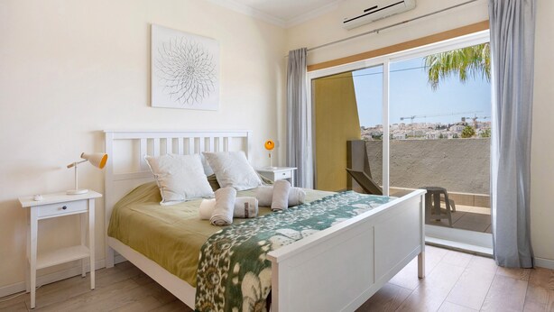 Un dormitorio con cama, mesita de noche, lámpara, ventana con vistas y cortina.