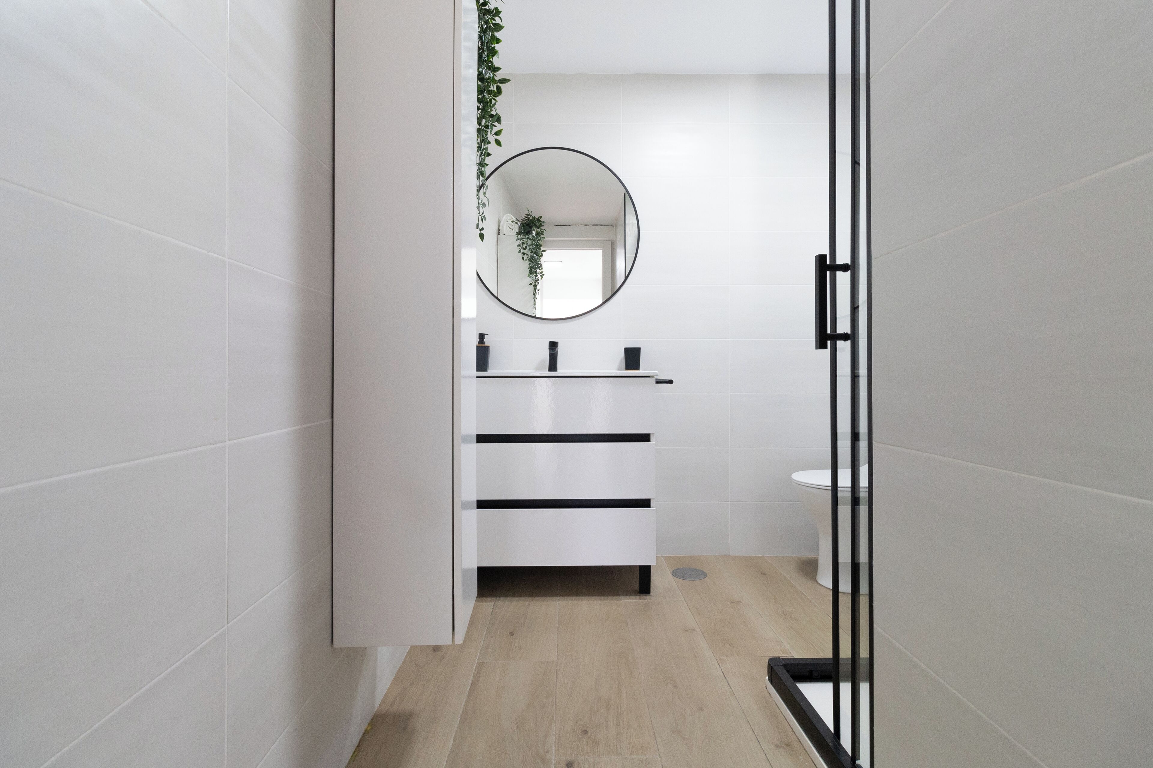 A modern bathroom with white tiles, a round mirror, a white sink, and a wooden floor.