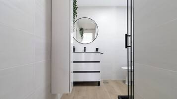 A modern bathroom with white tiles, a round mirror, a white sink, and a wooden floor.