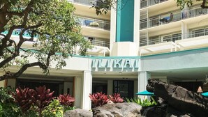 A hotel entrance with a rock waterfall feature, surrounded by tropical plants and a multi-story building with balconies.