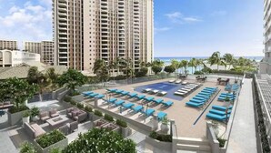 A resort pool area with multiple lounge chairs and a seating area, surrounded by palm trees and a large building in the background.
